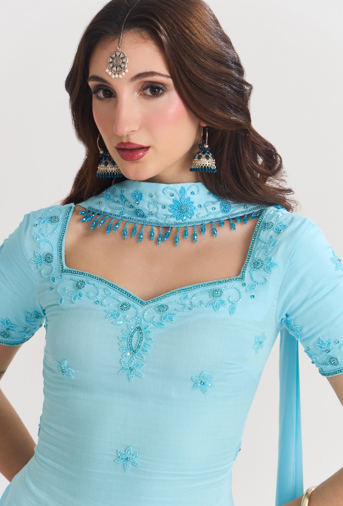 A woman wearing a light blue embellished traditional outfit with a high embroidered collar, short sleeves, and decorative embroidery on the chest and shoulders, photographed against a plain light background.